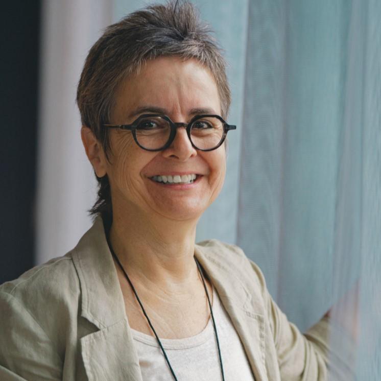 Porträtfoto von Stefanie Vonhoff. Sie trägt einen beigen Blazer und eine schwarze Brille und ist knapp bis zur Schulter zu sehen. Sie steht vor einem Fenster mit weißem Vorhang und lächelt in Richtung Kamera.  